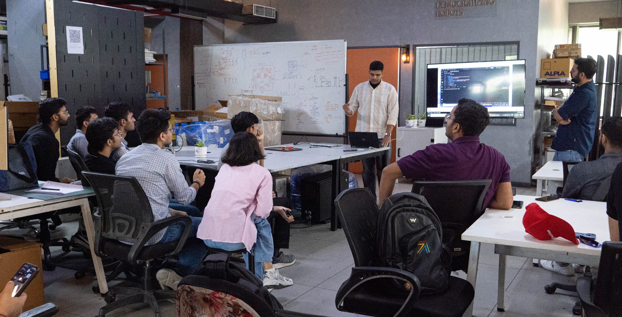 People sit in a meeting room watching a presenter stand beside a screen showing code. Tables, chairs, a whiteboard, and shelves fill the background in a dim indoor setting.