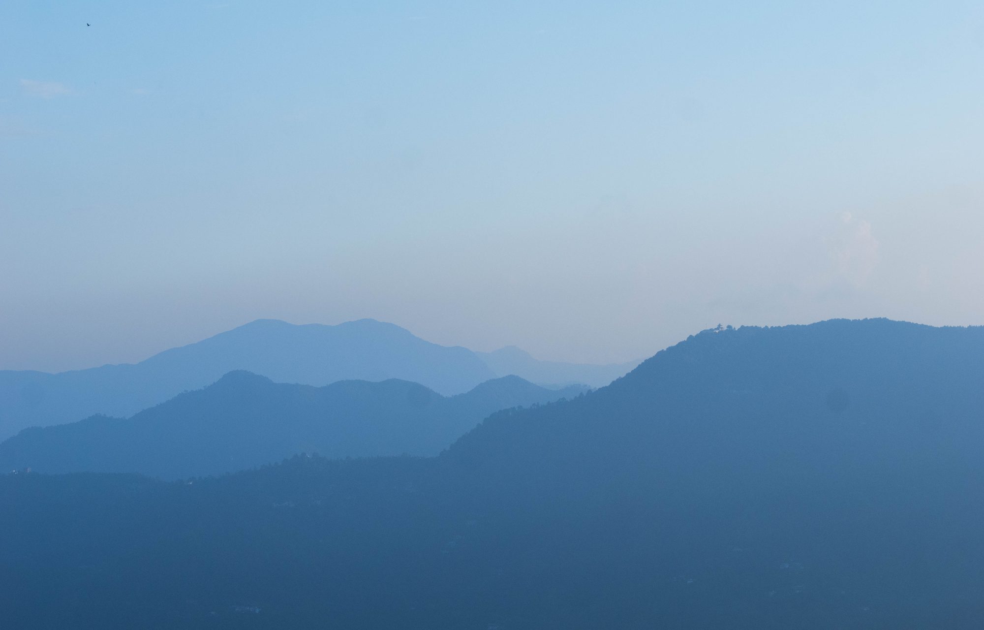 A hazy landscape of layered mountain ridges gradually fading into shades of blue beneath a pale, misty sky