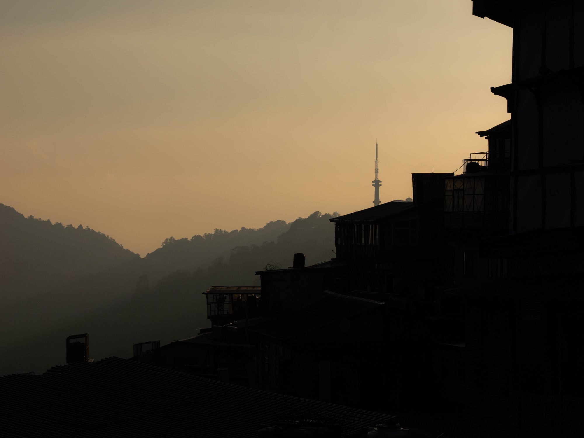 Dark silhouettes of buildings and hills frame a hazy golden sky, with a tall communications tower rising in the distance.