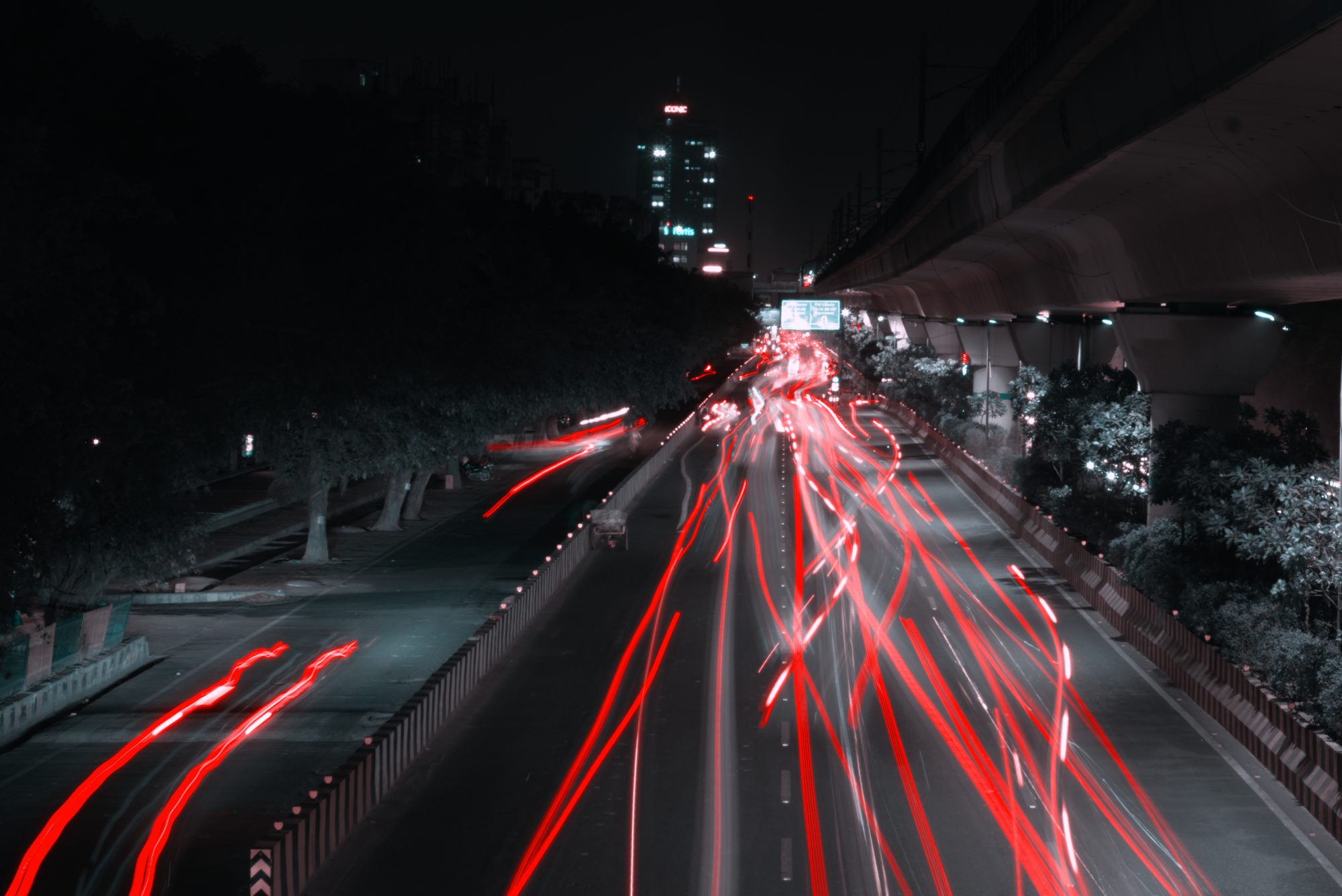 Long-exposure night view of a highway with bright light trails stretching into the distance, flanked by dark trees and an elevated structure on the right. A tall building glows faintly in the background.
